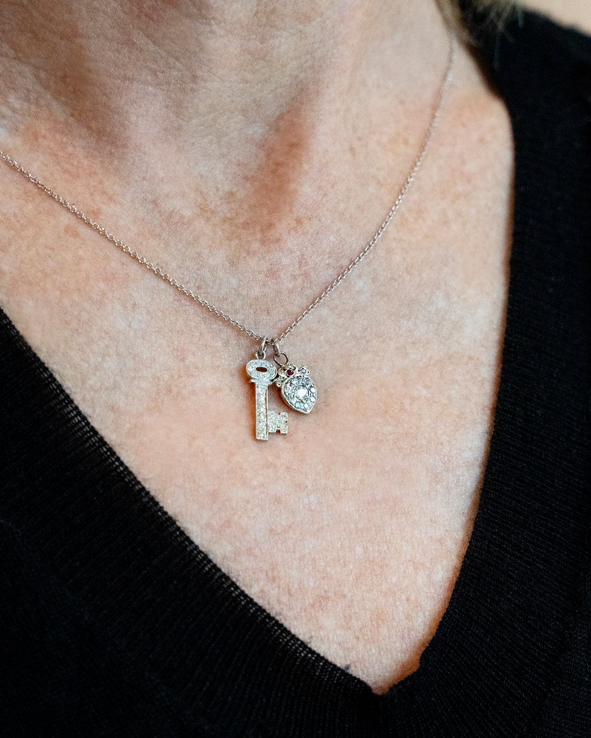 Diminutive Art Deco era ‘Key to my Heart’ charms on a platinum 18” cable link chain. A tiny heart crowned with a bow and ruby stone hanging with a matched key in platinum set with diamonds. Both pieces came from an early 20th-century charm bracelet. Continental European, 1930. Being worn on a model's necklace to show the scale of the necklace. Hanging on an 18" chain.