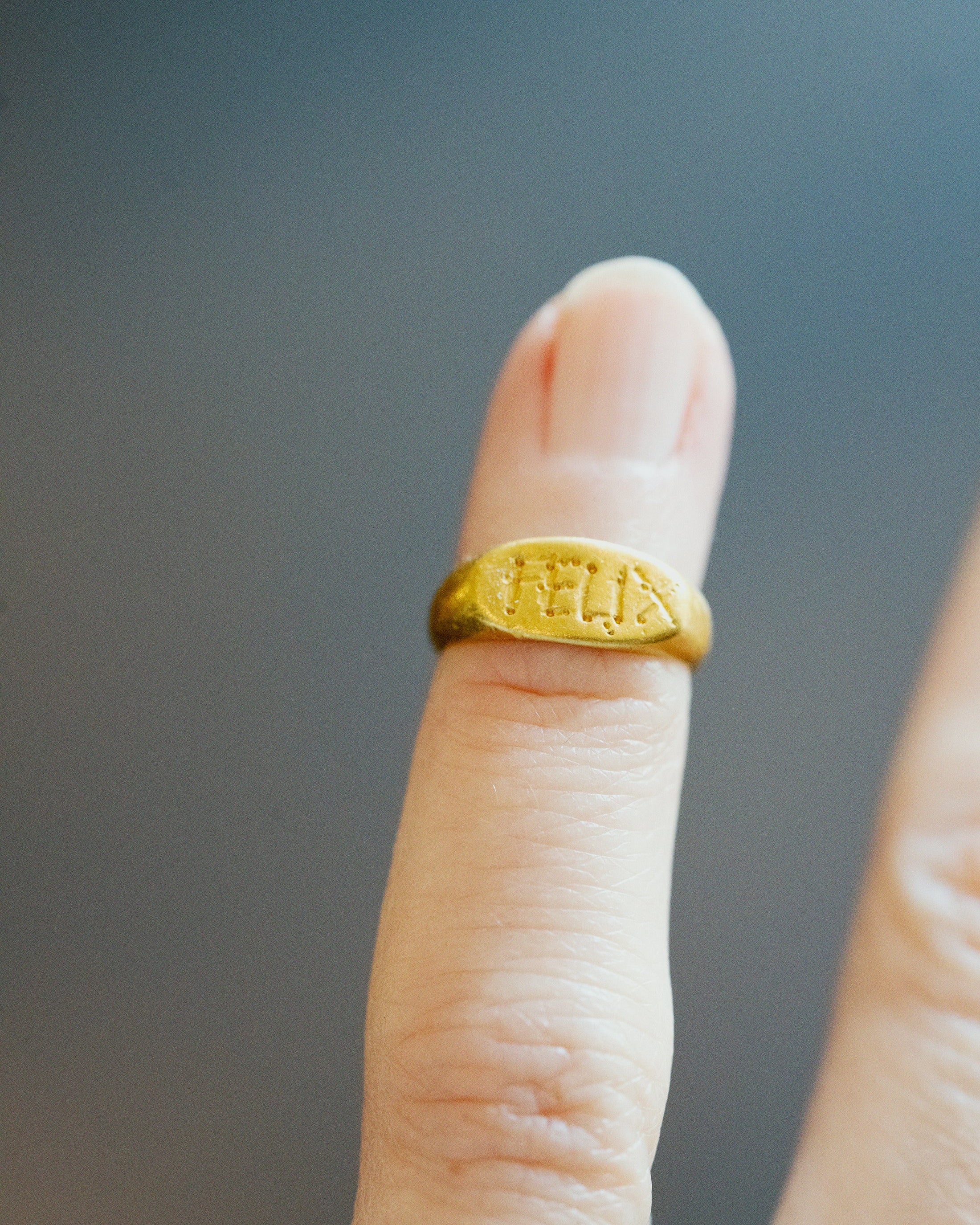 A close-up of a gold signet ring engraved with the name 'FELIX' on a finger, set against a dark gray background.