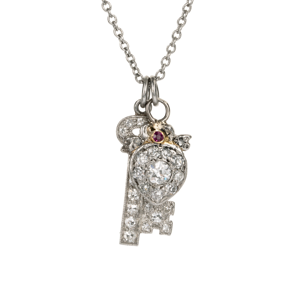 Diminutive Art Deco era ‘Key to my Heart’ charms on a platinum 18” cable link chain. A tiny heart crowned with a bow and ruby stone hanging with a matched key in platinum set with diamonds. Both pieces came from an early 20th-century charm bracelet. Continental European, 1930. Front view.