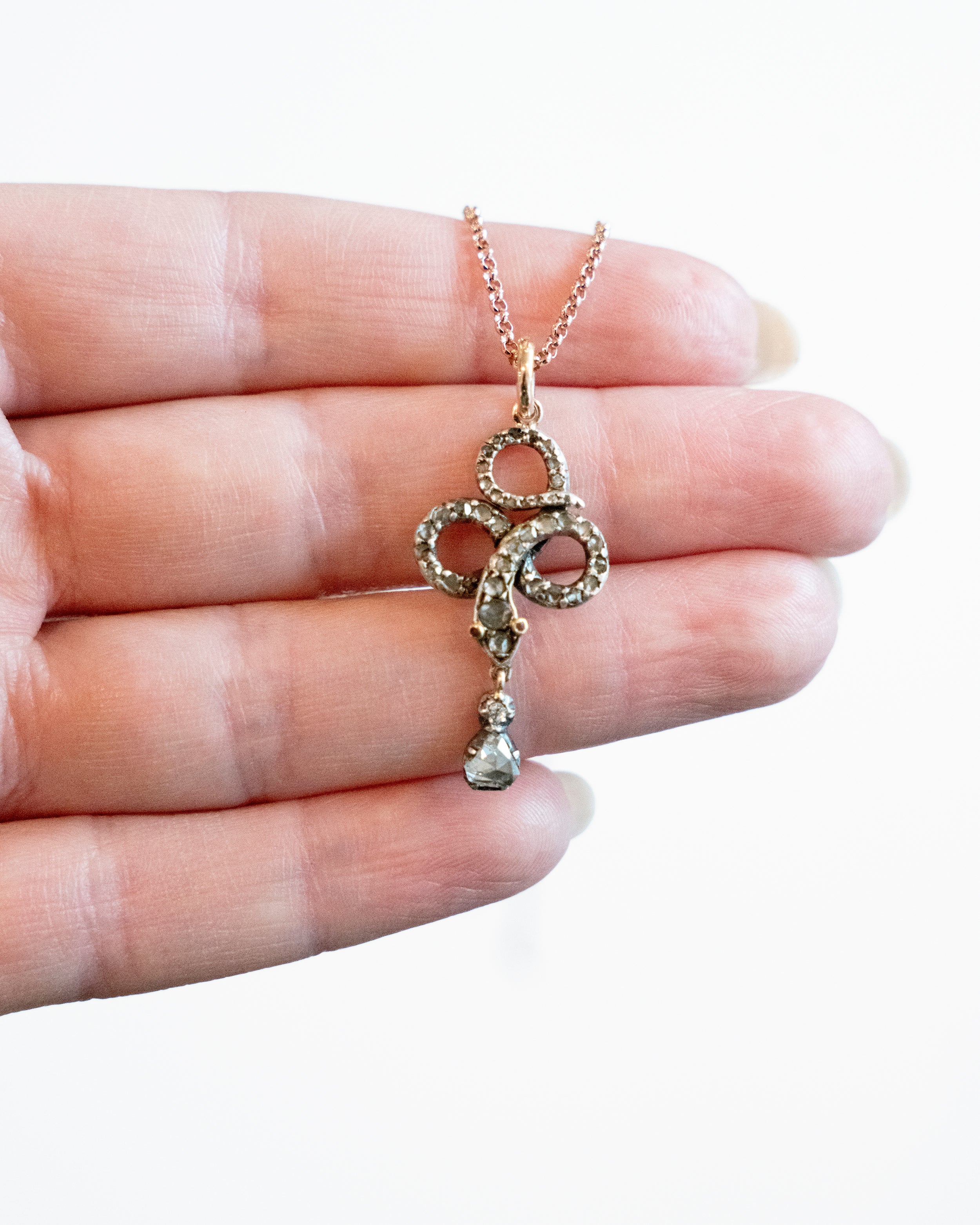 Rose cut diamonds are set  in silver topped gold in the form of a snake. The snake is coiled into a trefoil design with a tear drop shaped diamond drop. The detail extends to two tiny ruby eyes. The piece appears to have been converted into a pendant from a brooch or stick pin. England, Circa 1820. Being held in a hand for scale.