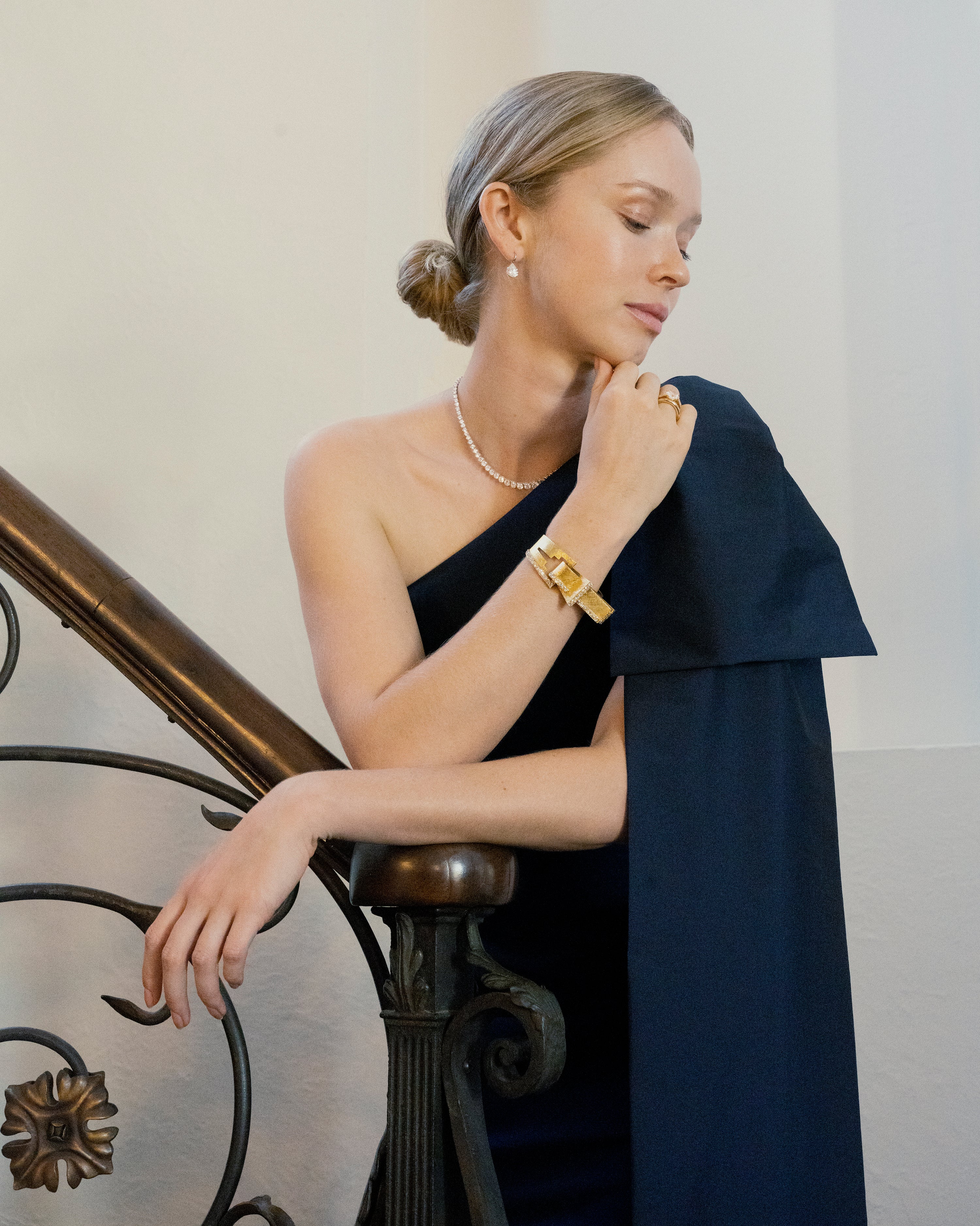 The American jewelry firm Trabert & Hoeffer, with the Parisian house Mauboussin, produced some of the most extraordinary jewels of its time. Famous for their illustrious Hollywood clientele. This chic 18k gold bangle has a florentine finish in an abstract buckle design with bright diamonds set in platinum.American in origin.Circa 1960. A model on stairs wearing a navy blue dress and wearing a diamond necklace, the bracelet and diamond earrings.