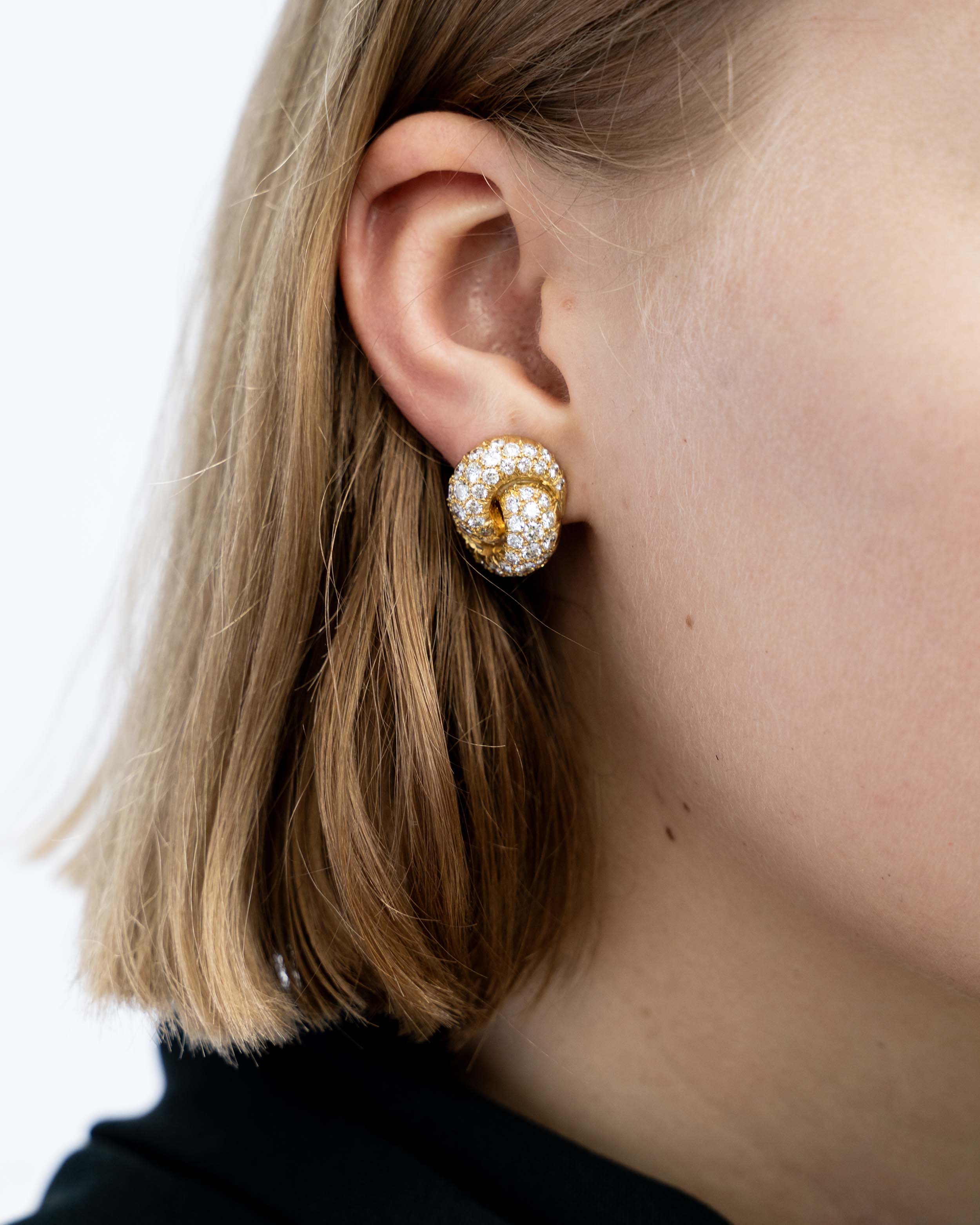 Vintage pave diamond knot earrings. Timeless ear clips in 18k gold. The intertwined loop design is set with approximately 4.50 carats in total diamond weight. The diamonds are estimated to be G to H in color, and VS1 to VS2 clarity. America, circa 1980. On a model.