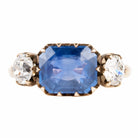 A three stone old mine cut diamond ring with a large blue sapphire center stone and two smaller old European cut side diamonds, all in a rose gold band.