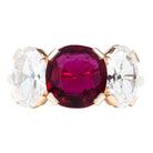 A ring featuring a red cushion-shaped ruby centerpiece flanked by two clear, old cut oval-shaped diamonds, all set in a simple, low profile band.