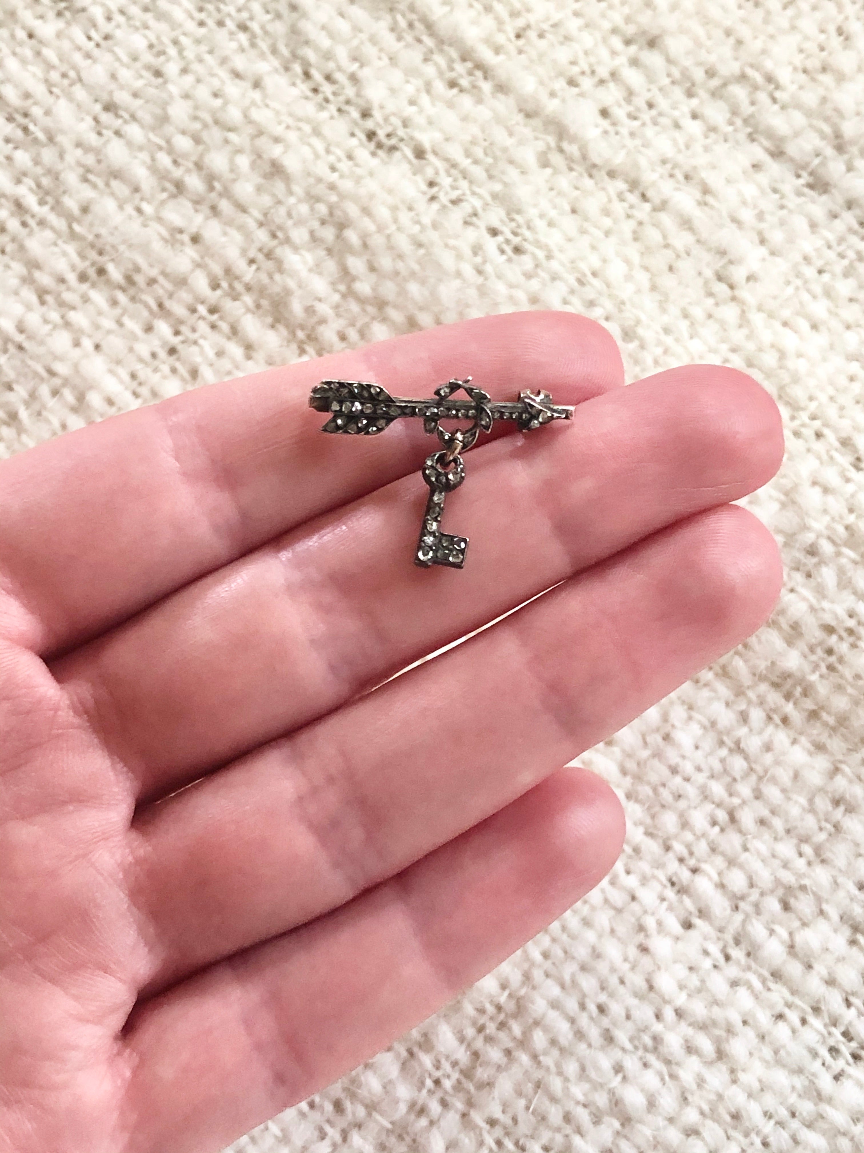Tiny Victorian era Rose cut diamond arrow brooch with the tiniest key hanging from a laurel wreath. Silver topped gold with original clasp fixture. English in Origin. Circa 1860. Close up shot on a hand for scale.