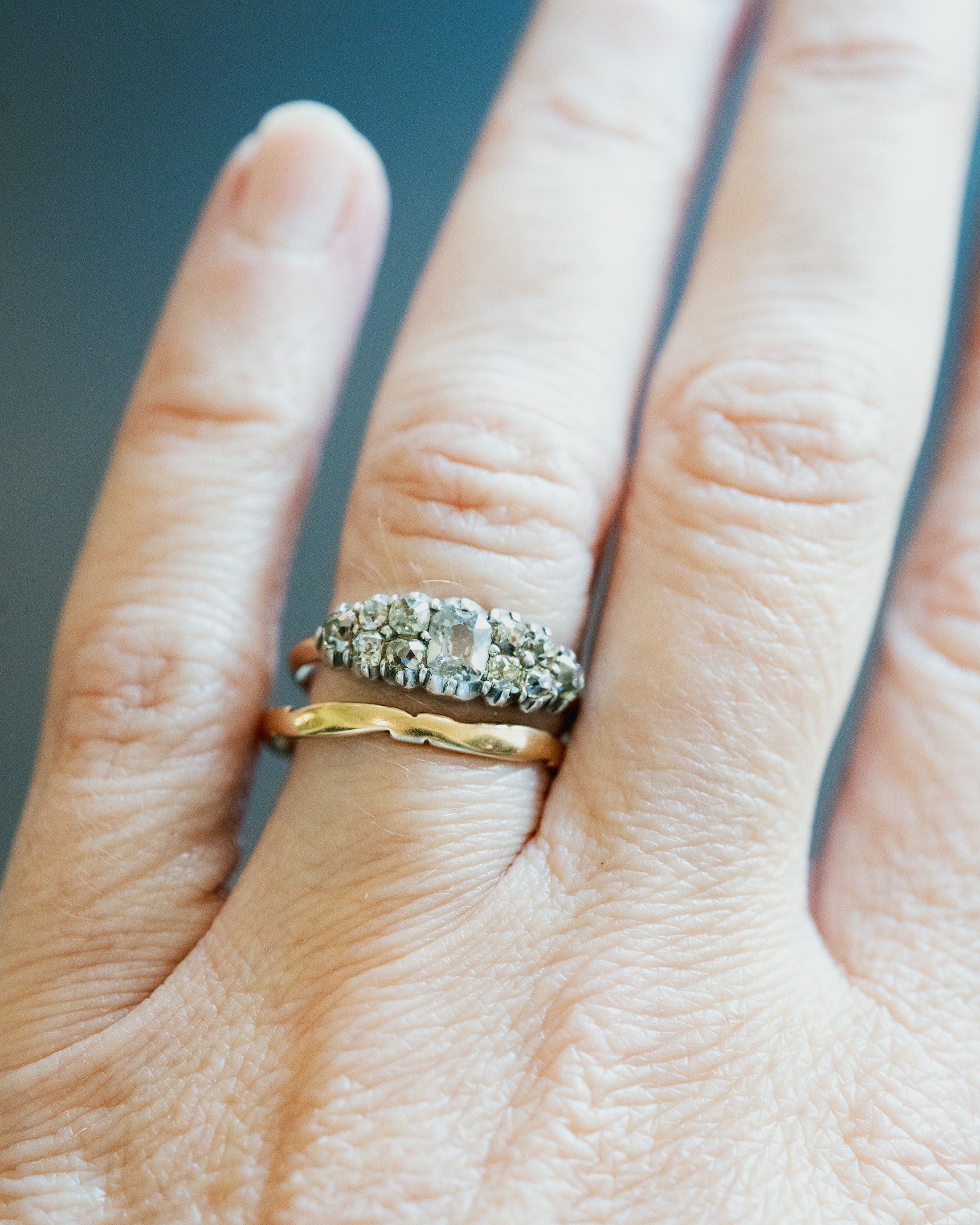 A close-up of a hand wearing three rings: a silver-toned ring with a central clear marquise-cut stone and smaller surrounding stones, a gold band, and a textured silver ring.