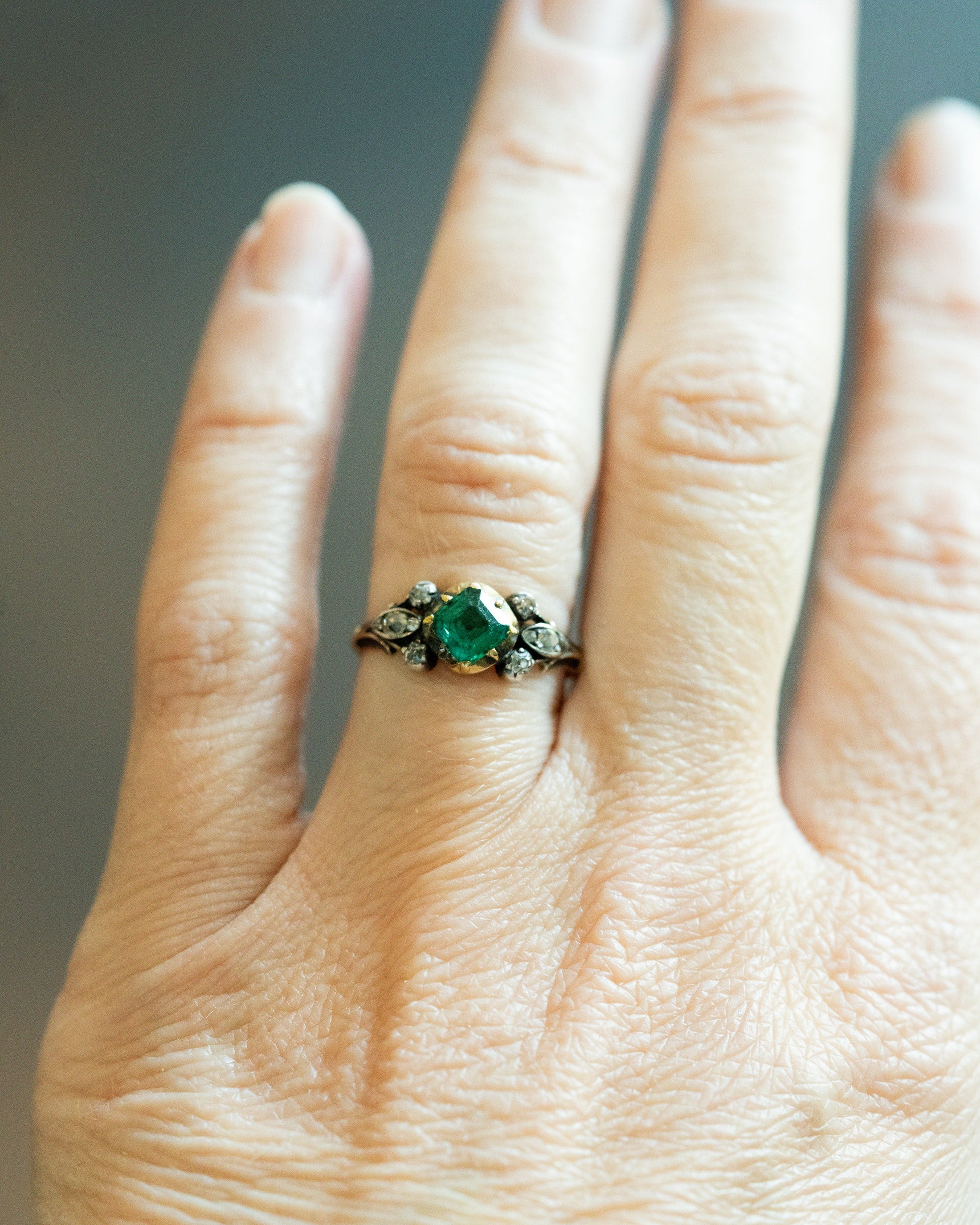 A close-up of a hand wearing an antique-style ring with a central green gemstone and small clear stones on a dark band.