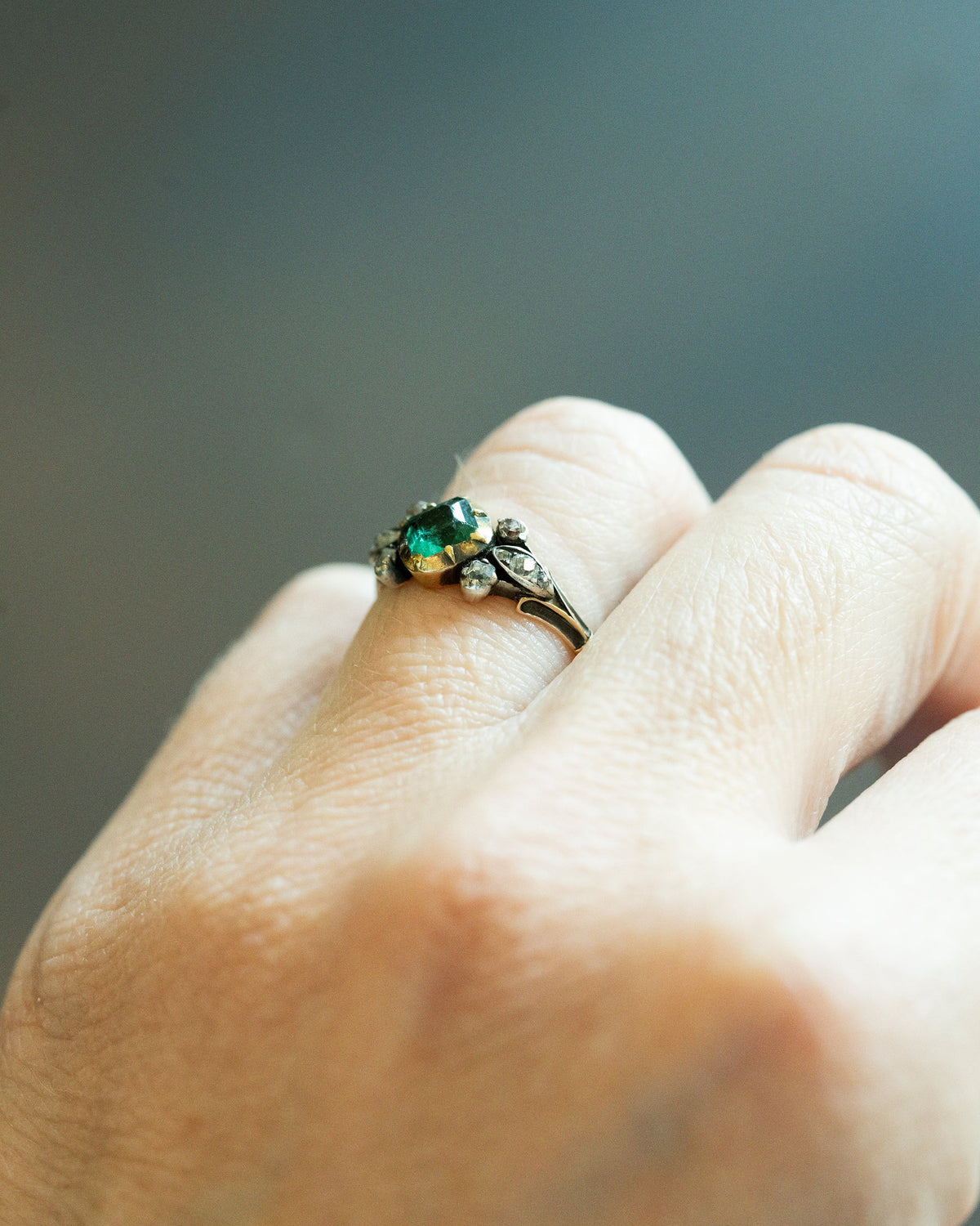 A close-up of a hand wearing a vintage-style ring with a green oval stone and small clear stones on a dark metal band, set against a blurred gray background.