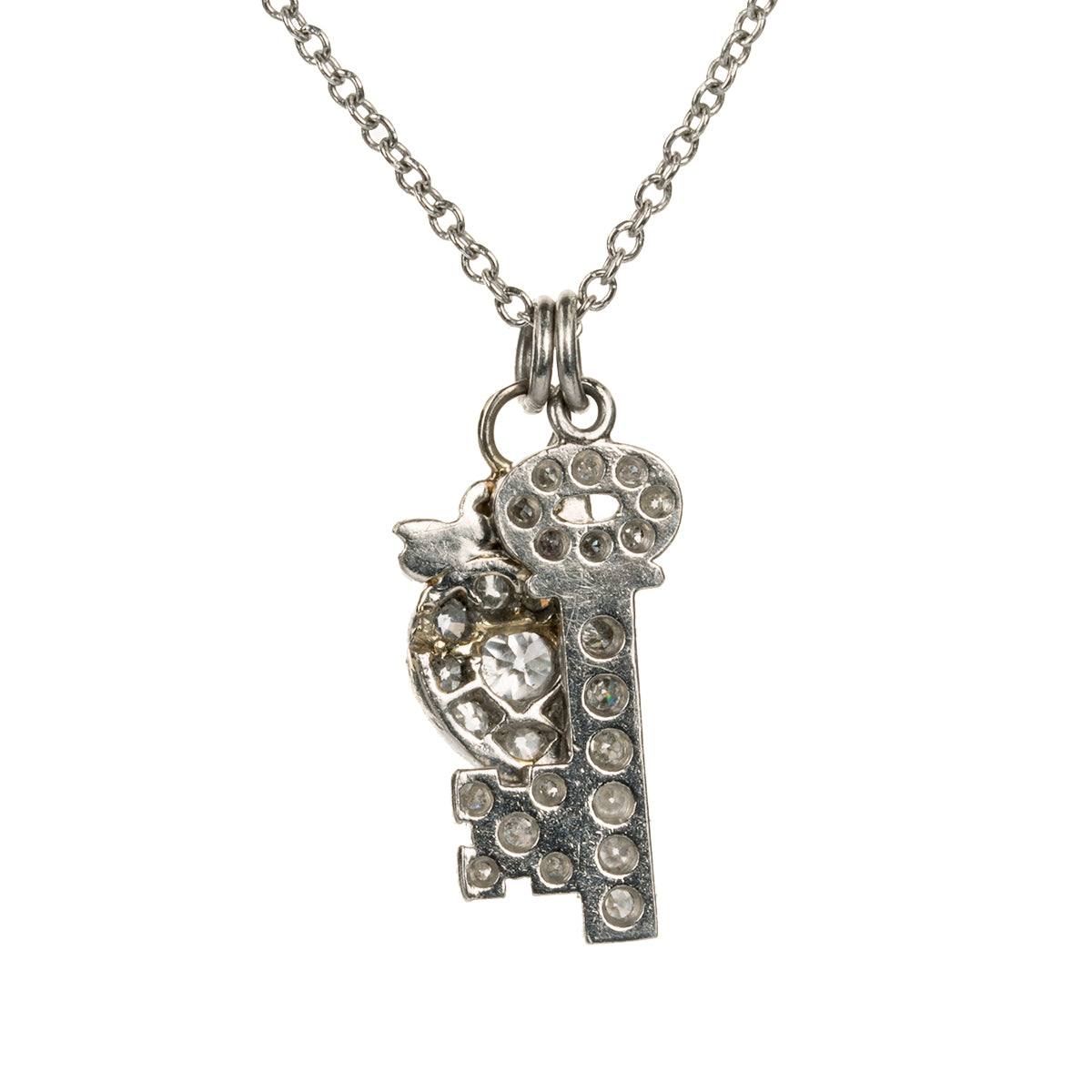 Diminutive Art Deco era ‘Key to my Heart’ charms on a platinum 18” cable link chain. A tiny heart crowned with a bow and ruby stone hanging with a matched key in platinum set with diamonds. Both pieces came from an early 20th-century charm bracelet. Continental European, 1930. Close up of the back side of the charms. 