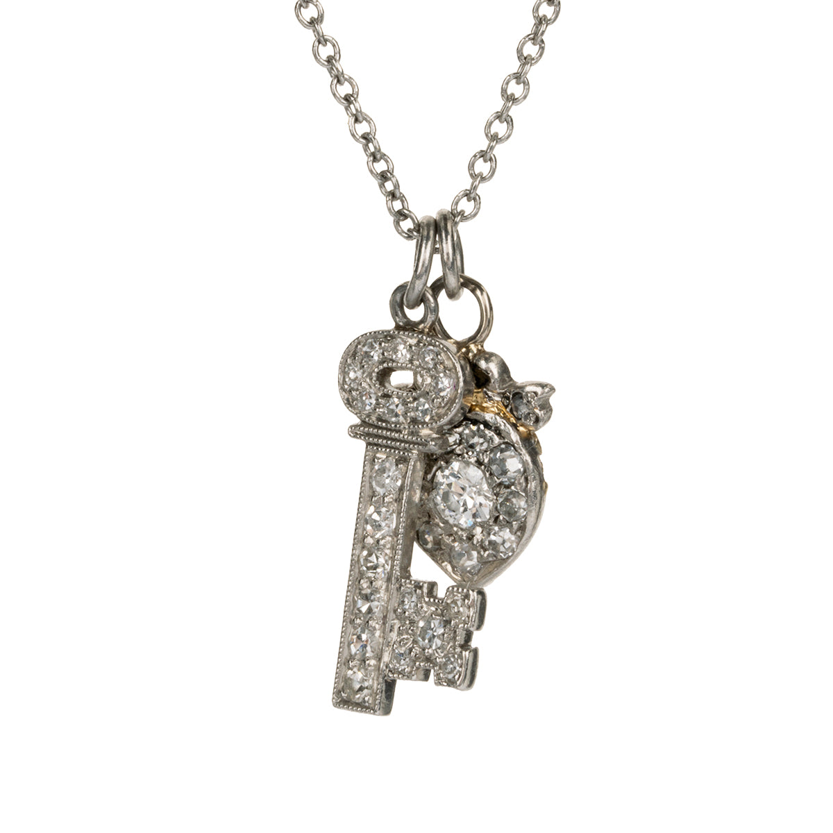 Diminutive Art Deco era ‘Key to my Heart’ charms on a platinum 18” cable link chain. A tiny heart crowned with a bow and ruby stone hanging with a matched key in platinum set with diamonds. Both pieces came from an early 20th-century charm bracelet. Continental European, 1930. Close up view of the charms. 