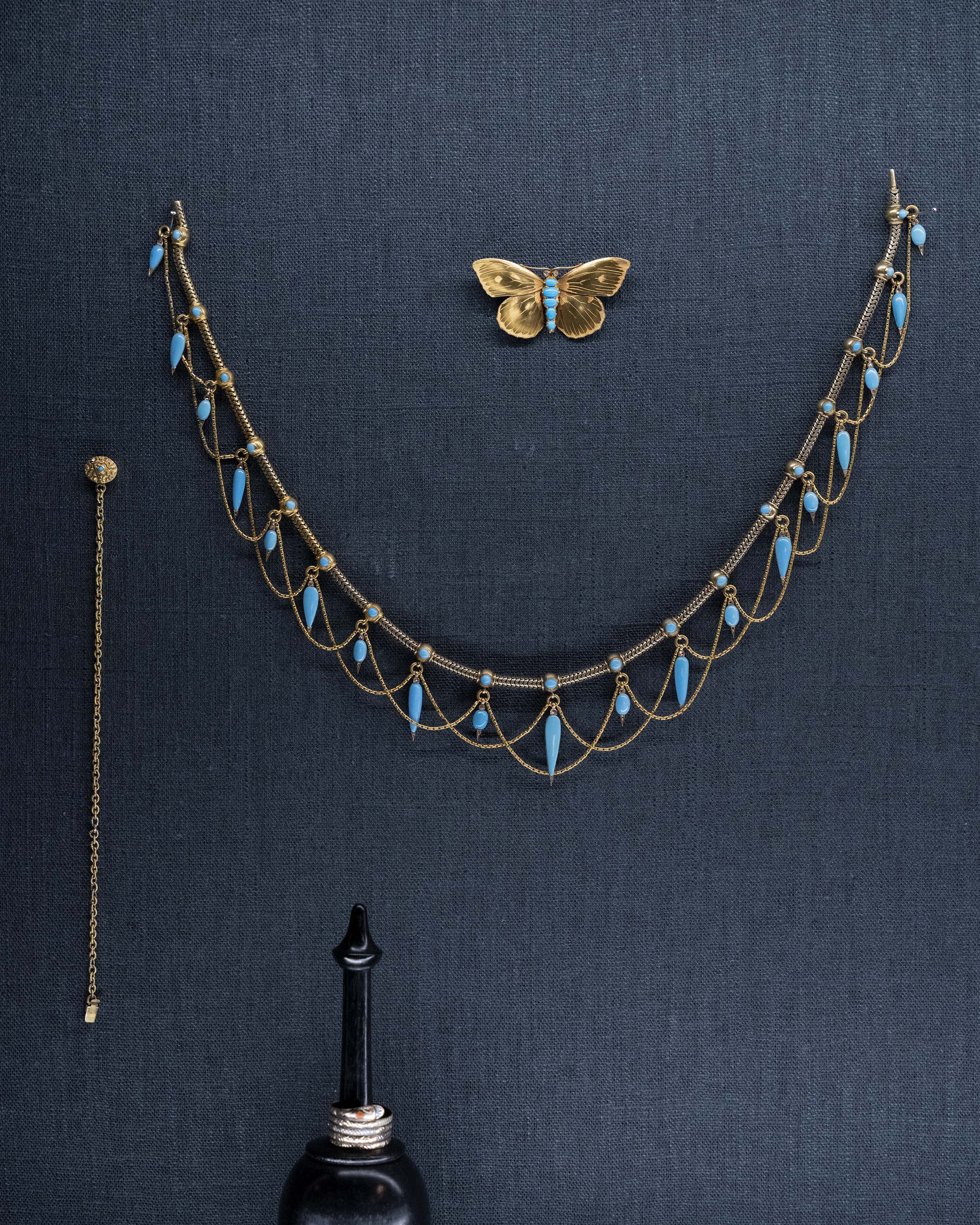This is a rare example of an elaborate 19th-century collar necklace. Natural Persian turquoise stones are cut in the shapes of amphoras, with rose-cut diamonds set at the sharp tip of every setting. The necklace is made of 18k gold and was made in England, circa 1860. The necklace on display with a butterfly brooch and a turquoise chain bracelet.