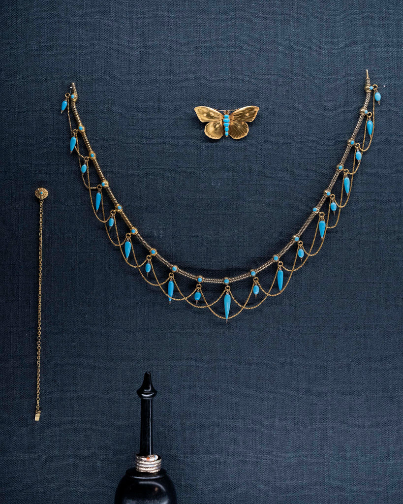 This is a rare example of an elaborate 19th-century collar necklace. Natural Persian turquoise stones are cut in the shapes of amphoras, with rose-cut diamonds set at the sharp tip of every setting. The necklace is made of 18k gold and was made in England, circa 1860. The necklace on display with a butterfly brooch and a turquoise chain bracelet.