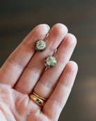 A close-up of a person's hand holding a pair of white paste earringss, with two gold rings on the fingers, against a dark blurred background.