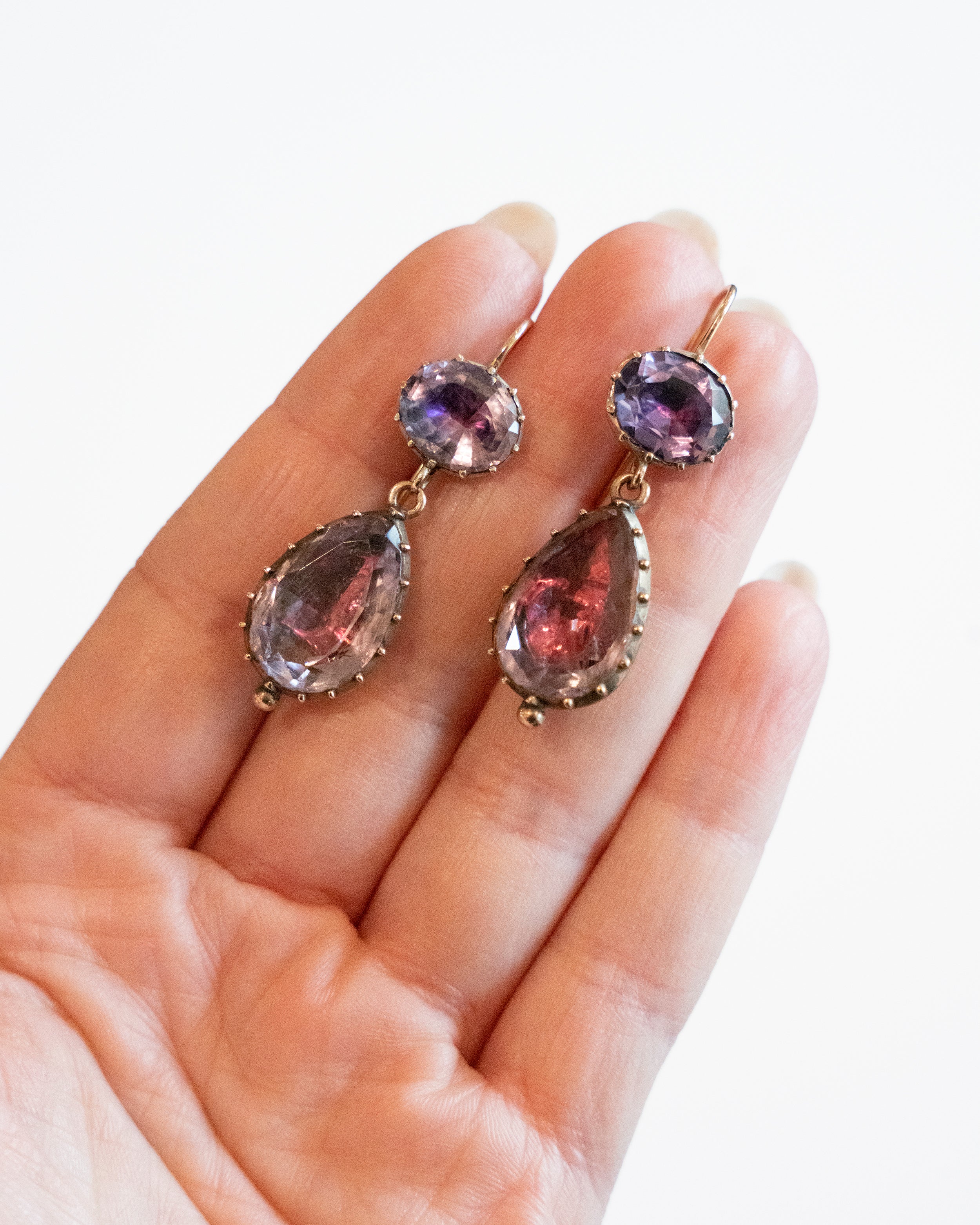Lovely early 19th century earrings set in 15k gold. Cushion and pear shaped amethyst stones set in closed-back collet settings. The stones are a pale purple, with varied shades of purple and pink coming from the foil behind the stones. The coloring is very unique. English in origin. Circa 1800. Hand holding earrings for scale. 